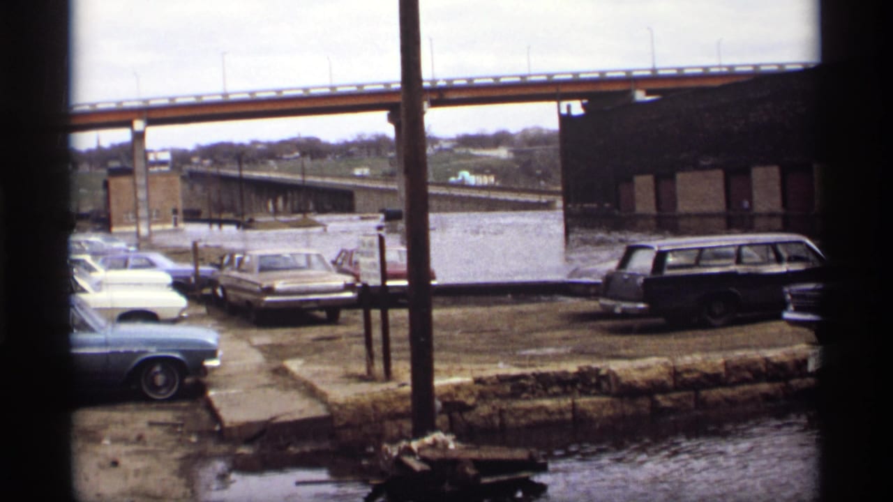 Flooded lot with stranded cars, bridge span, brick building, and utility poles.