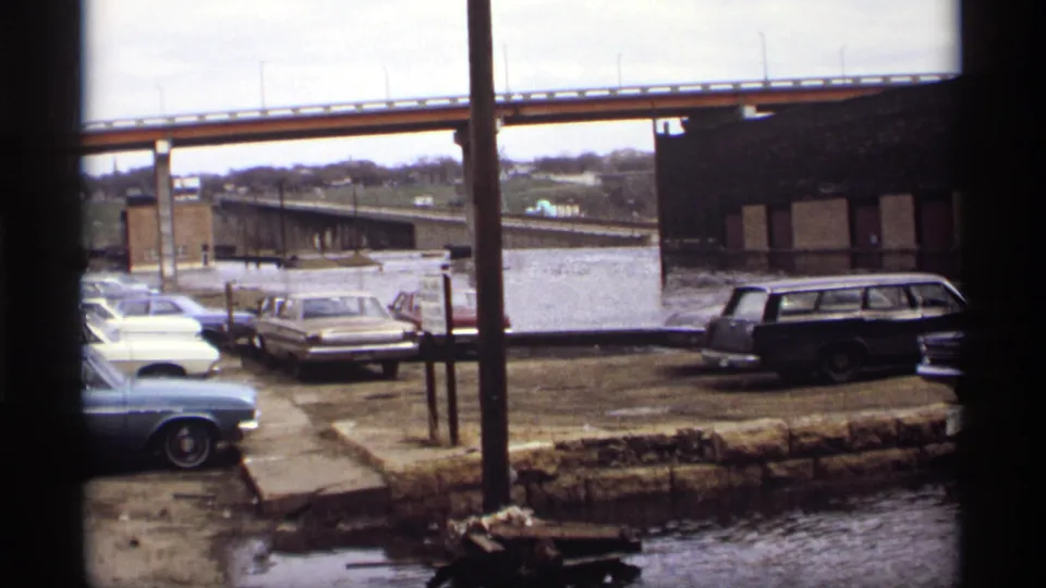 Saint Paul 1969 home-movie frame of floodwater around parked vehicles beside a bridge and brick building.