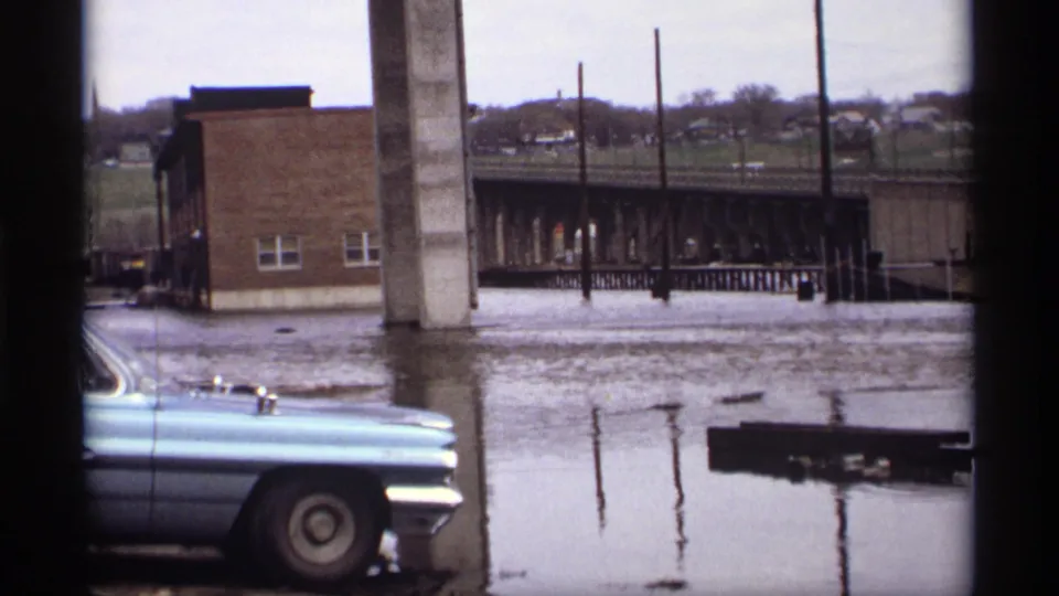 Saint Paul 1969 flood view with a blue sedan partly at frame edge, standing water, bridge support, and brick building.