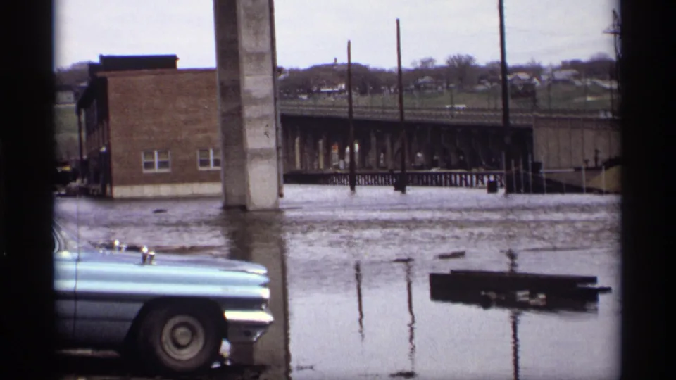 Saint Paul 1969 frame showing floodwater beside a blue sedan with bridge supports and a brick building in the background.