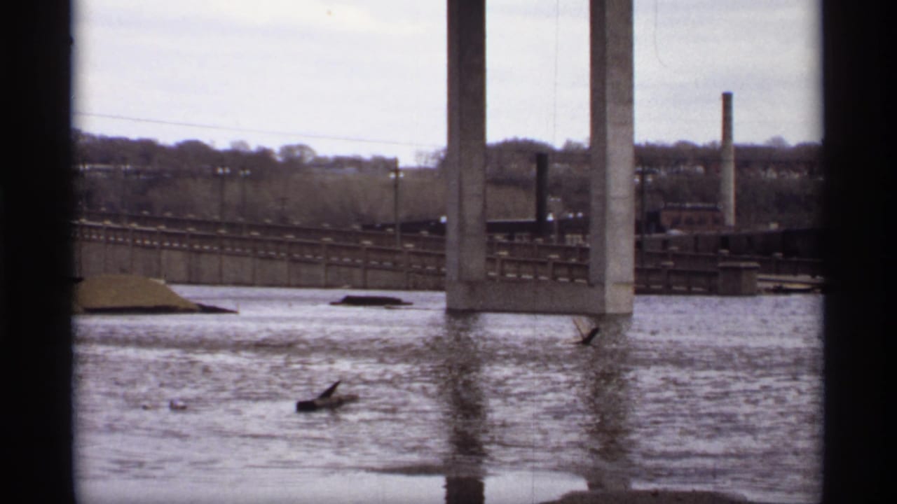 1969 Saint Paul 8mm frame showing floodwater under bridge piers, with rail lines, industrial stacks, and a distant treeline.