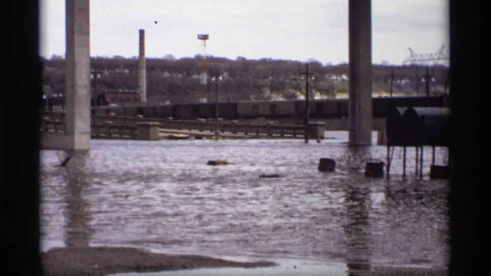 1969 Saint Paul footage frame with high water below bridge columns, rail cars in the background, chimneys, and small debris on the river.