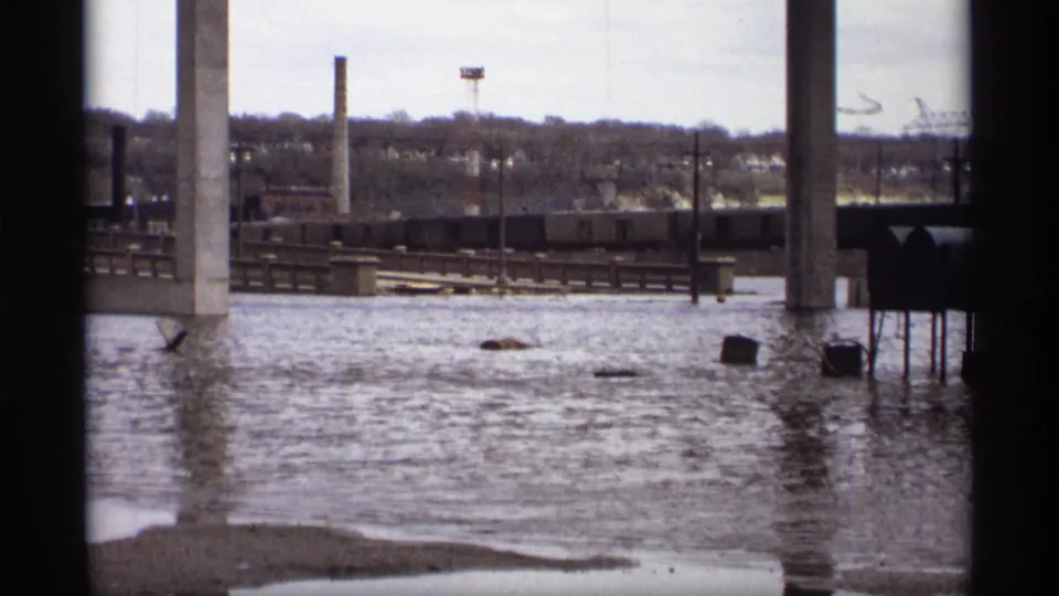 Saint Paul 1969 8mm frame looking across floodwater toward rail cars, bridge supports, industrial stacks, and a wooded ridge.