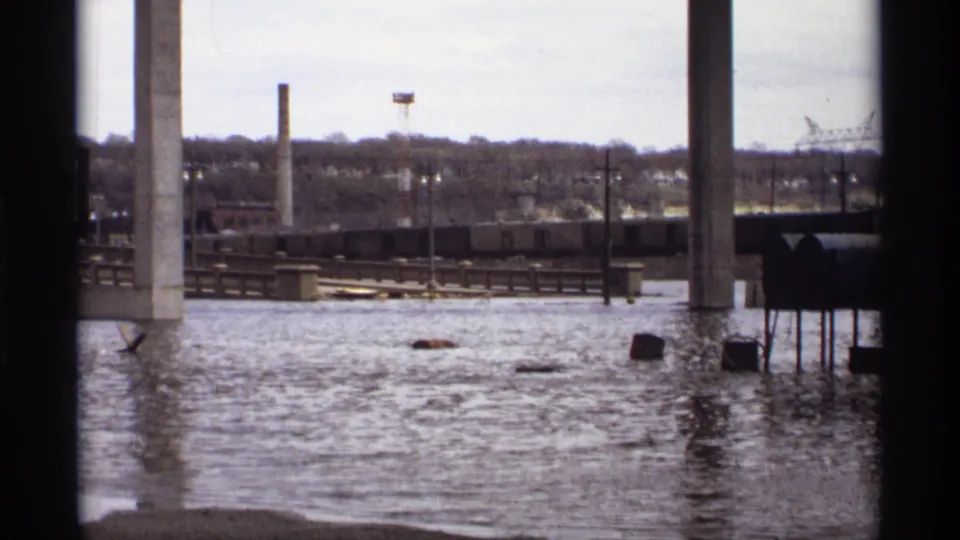 1969 Saint Paul home-movie frame showing floodwater around bridge piers, low riverfront structures, rail cars, and industrial chimneys.