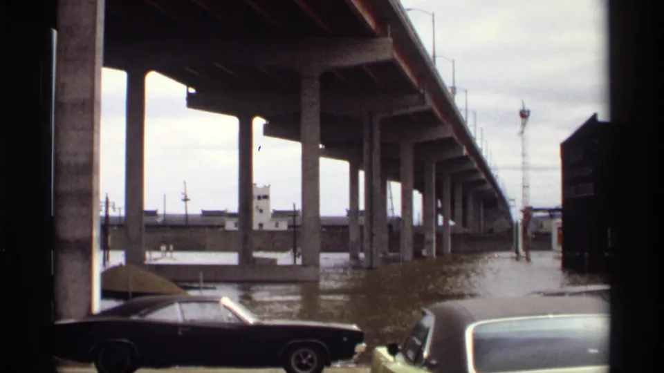 1969 Saint Paul flood scene showing muddy water under a concrete bridge, with period cars parked near the flooded riverfront.