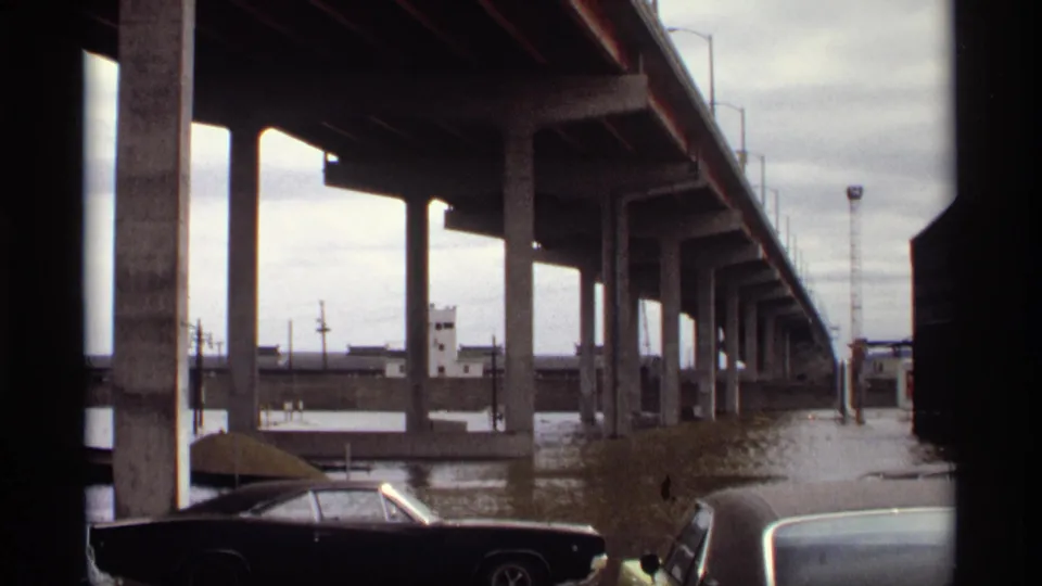 8mm view of a 1969 Saint Paul flood, with bridge pillars, murky water, and parked late-1960s cars along the riverfront.
