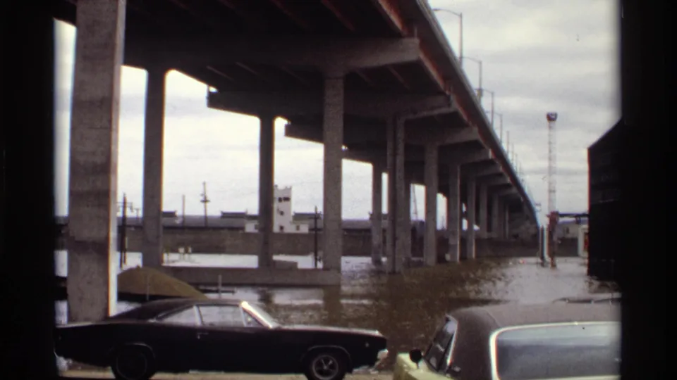 Flooded Saint Paul riverfront in 1969, showing concrete bridge supports, cloudy sky, industrial structures, and parked period cars.