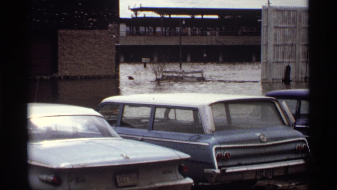 1969 Saint Paul home-movie frame showing floodwater around parked cars beside brick structures.