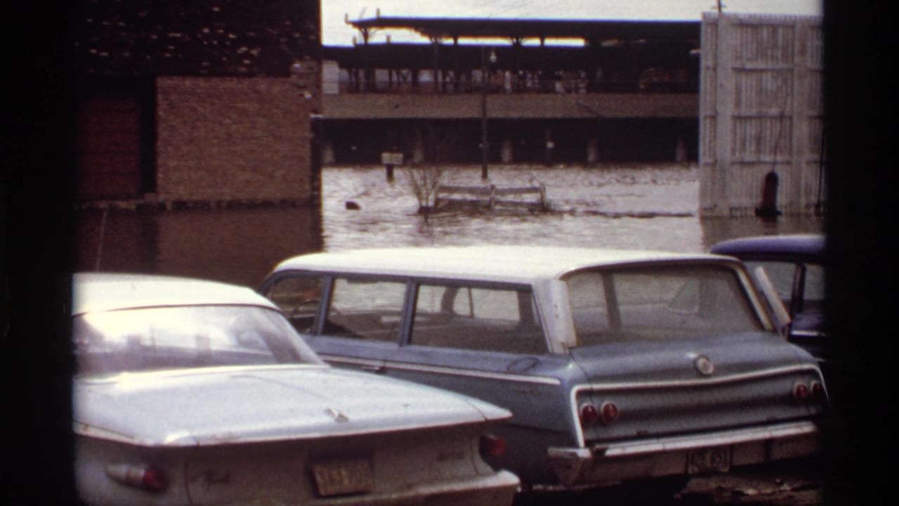 Parked cars sit in floodwater beside brick buildings and an open gate.