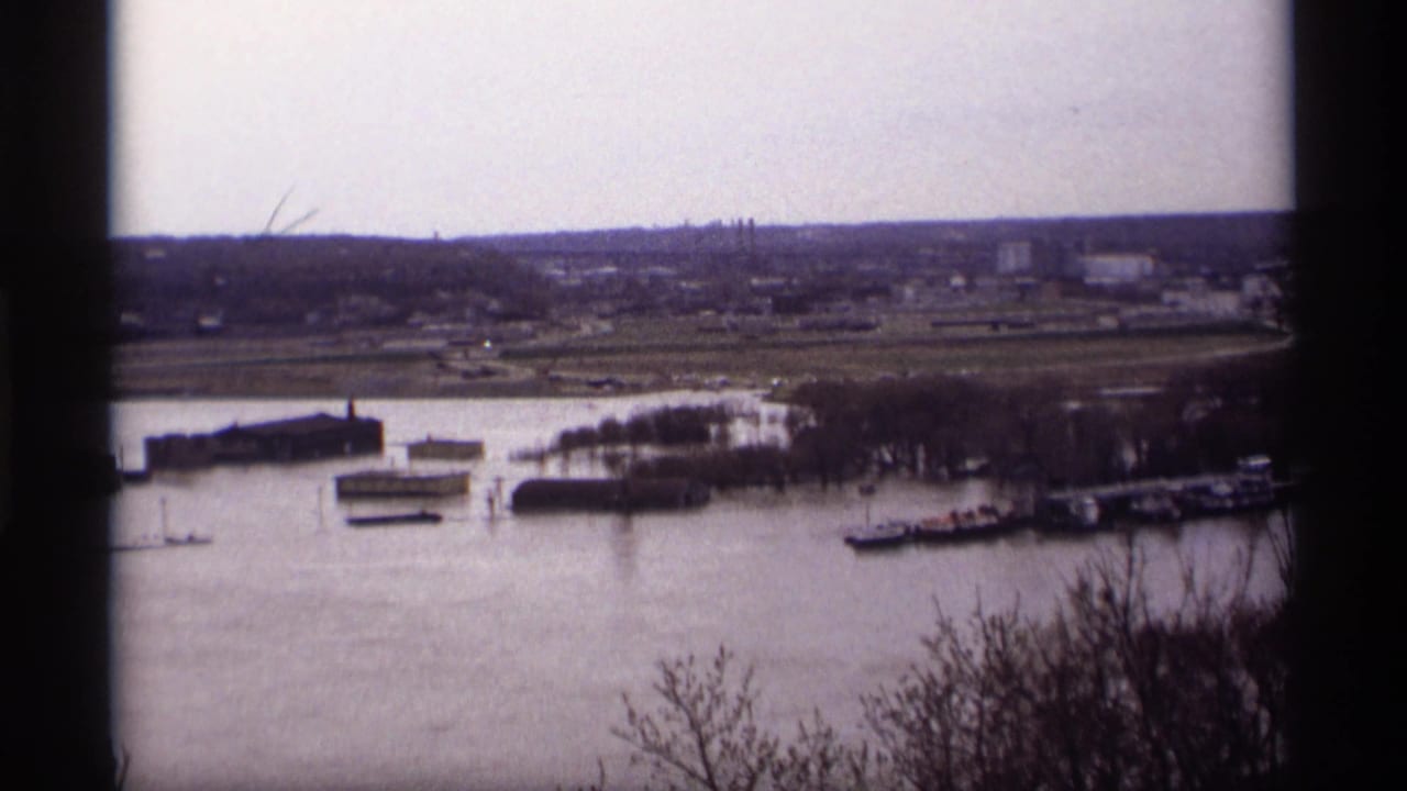 High-angle view of a flooded river with docks, buildings, bare trees, and distant industrial shoreline.
