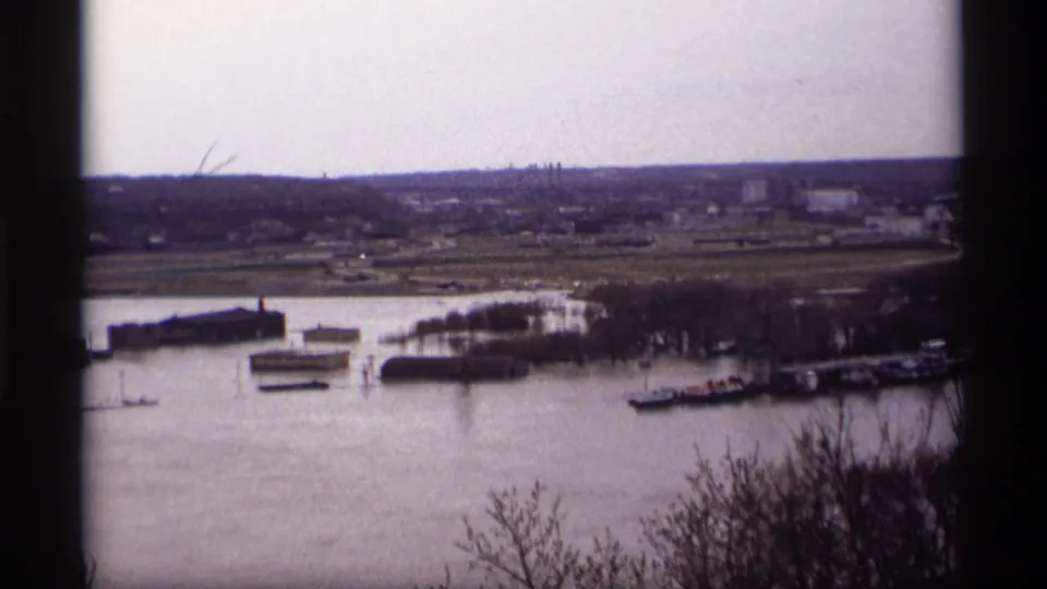 1969 Saint Paul 8mm frame showing floodwater around riverfront docks and low buildings, with bare trees and an industrial shore in the distance.