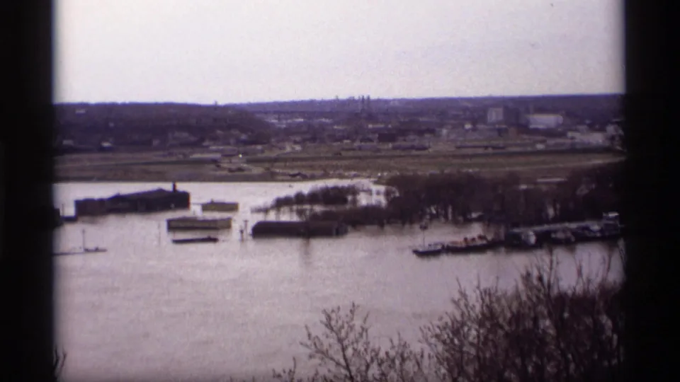 High-angle 1969 Saint Paul view of a swollen river covering docks near low waterside buildings, with leafless trees and distant structures beyond.