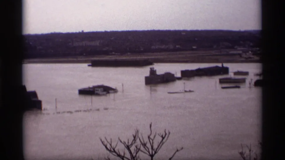 1969 Saint Paul flood scene with water spread across the riverfront, a small platform or dock, low buildings, and bare foreground branches.
