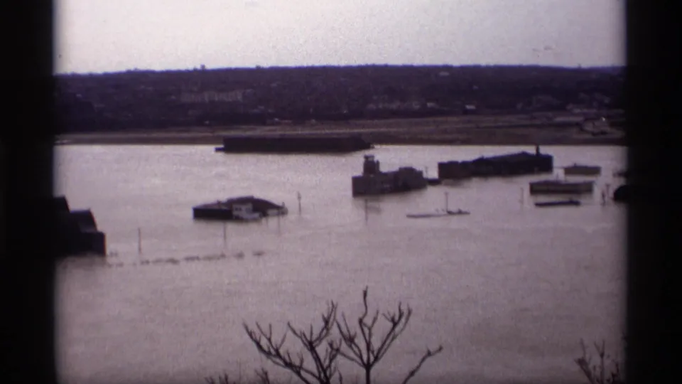 Final 1969 Saint Paul frame showing floodwater across the riverfront, with low structures, dock sections, bare branches, and a distant shore.