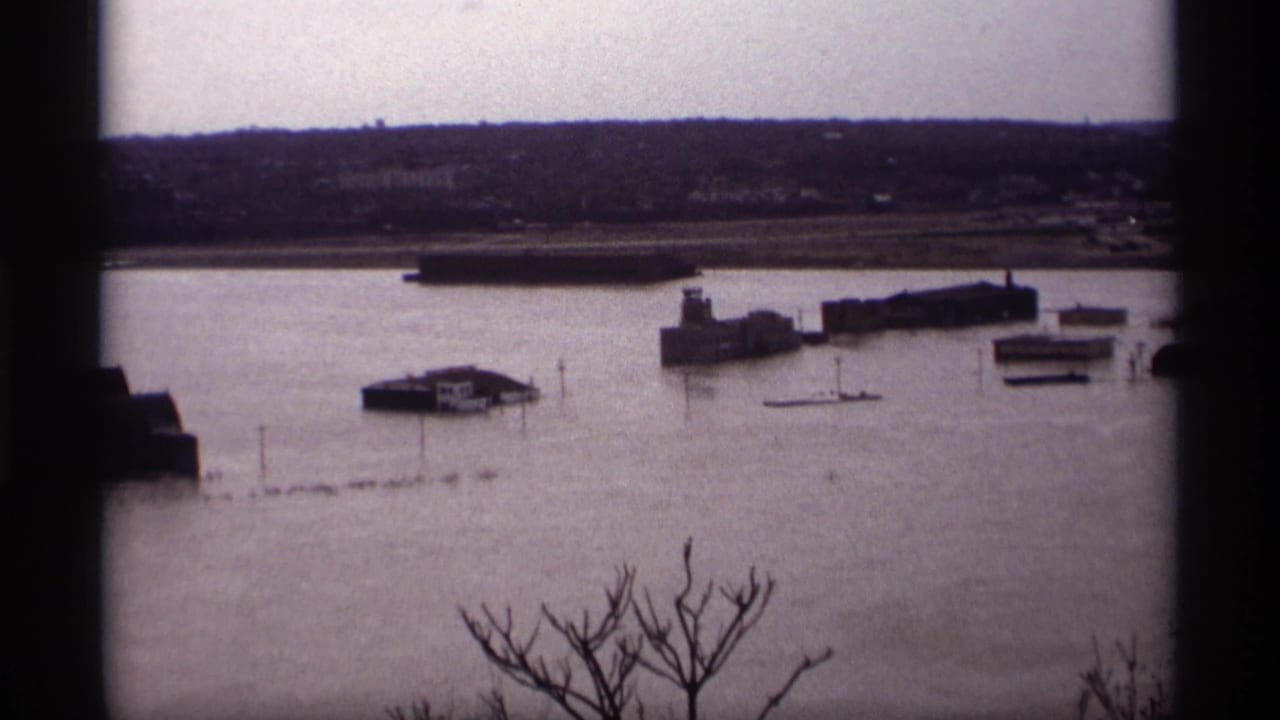 Flooded waterfront with industrial buildings, small structures, distant shore, bare branches, and dark frame borders.