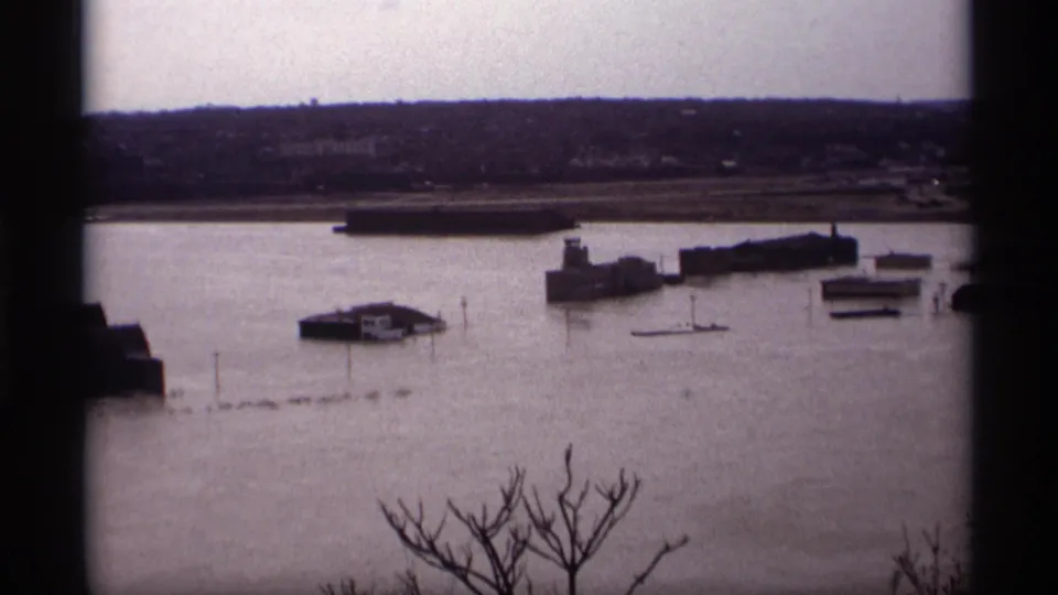 1969 Saint Paul 8mm frame showing floodwater around industrial waterfront buildings, with bare branches in the foreground and a distant shoreline.