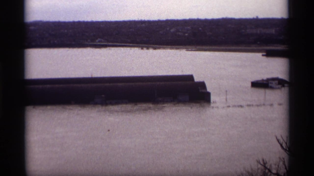Wide flooded riverfront with long industrial sheds, distant bank, pale sky, and dark vertical film border.