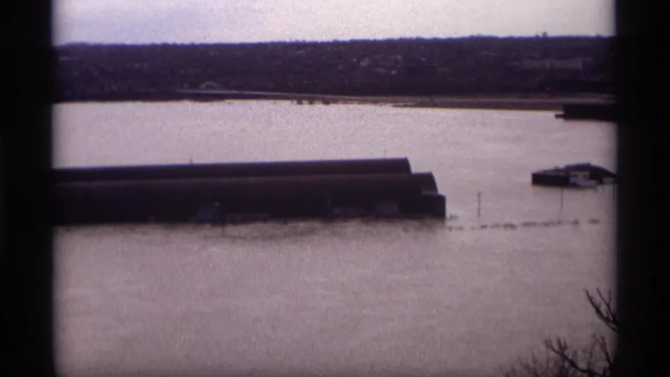 1969 Saint Paul home-movie frame looking over floodwater toward a long waterfront building, a small riverside structure, and a distant shore.