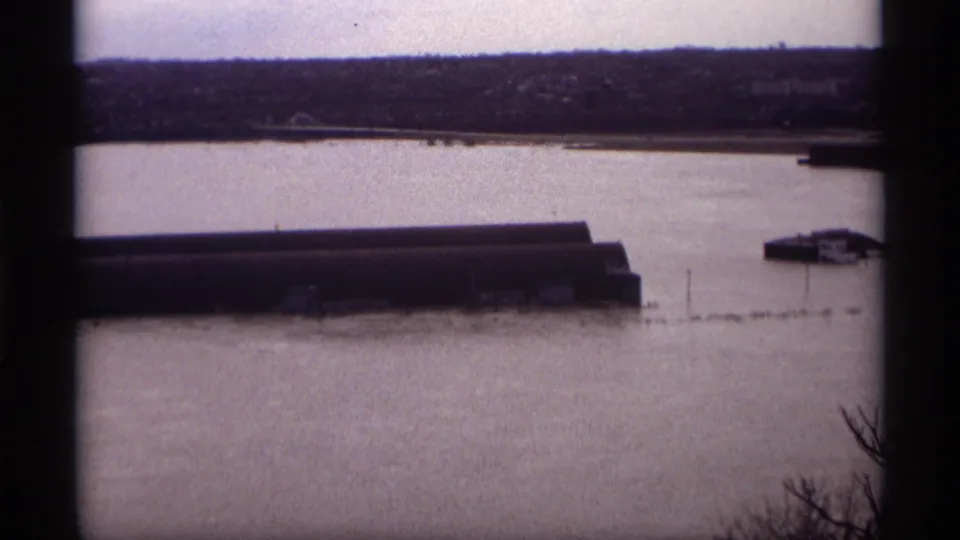 Saint Paul 1969 8mm frame showing a flooded industrial waterfront, with a long dark building, small riverside structure, and bare branches at the edge.