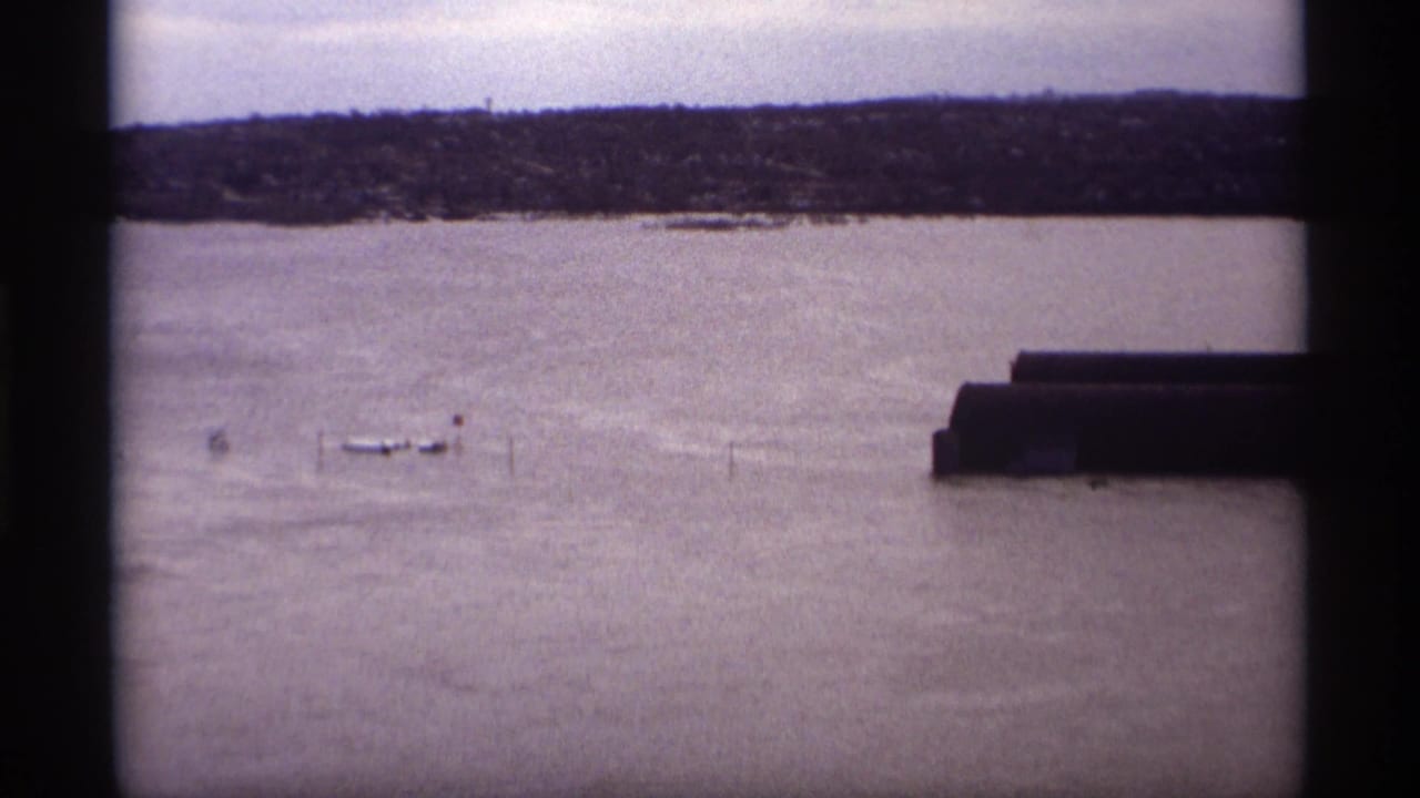 Flooded river seen through dark film-frame borders with boats, posts, distant shoreline, and overcast sky.
