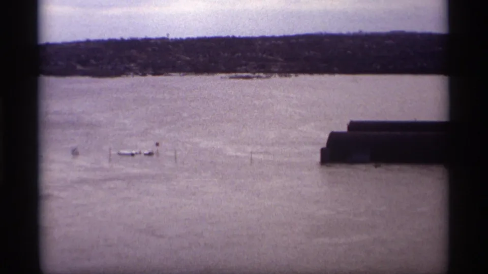 1969 8mm view of floodwater in Saint Paul, Minnesota, with small boats, submerged posts, a dark structure, and a distant treeline.