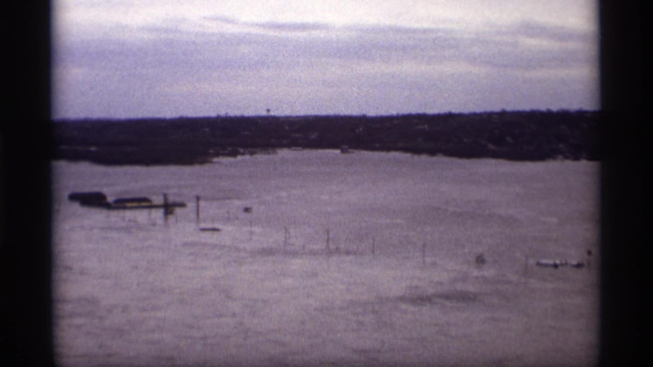 High water covers a lakeshore with partly submerged docks and distant buildings under a cloudy sky.