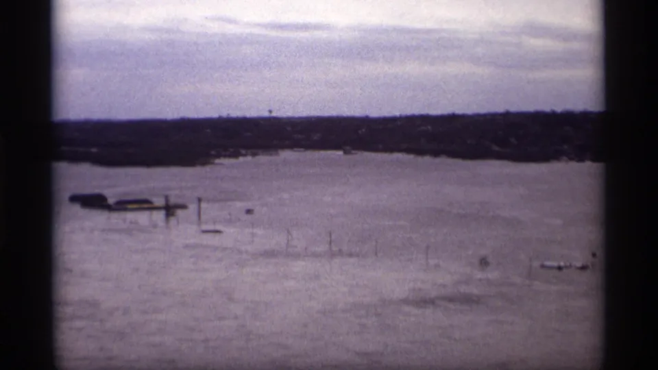 1969 Saint Paul 8mm frame showing a flooded lake, submerged dock structures, bare branches, and distant shoreline buildings.