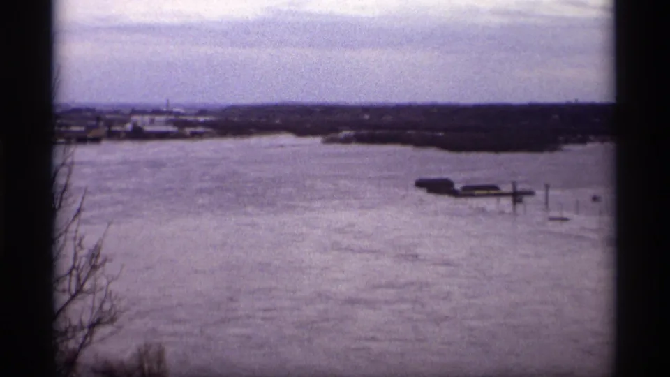 Wide 1969 Saint Paul lake view with elevated water, dock posts, low structures, bare branches, and a cloudy horizon.