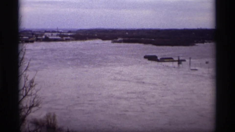 Saint Paul home-movie frame from 1969 showing high water around dock structures with bare shoreline branches in the foreground.