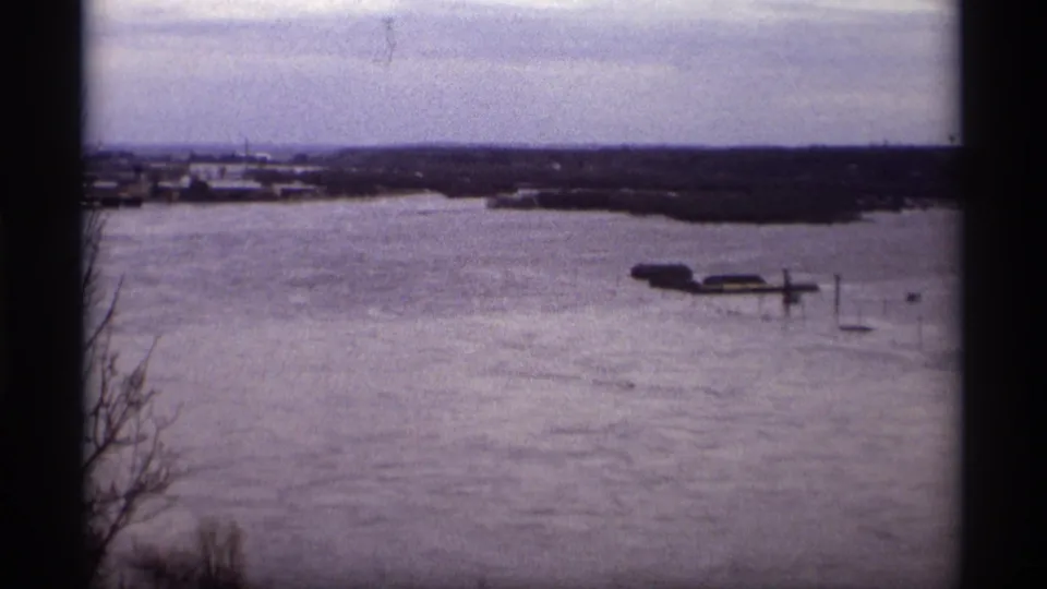 1969 Saint Paul 8mm frame of a flooded lakeshore, with dock structures in high water and distant buildings beneath an overcast sky.