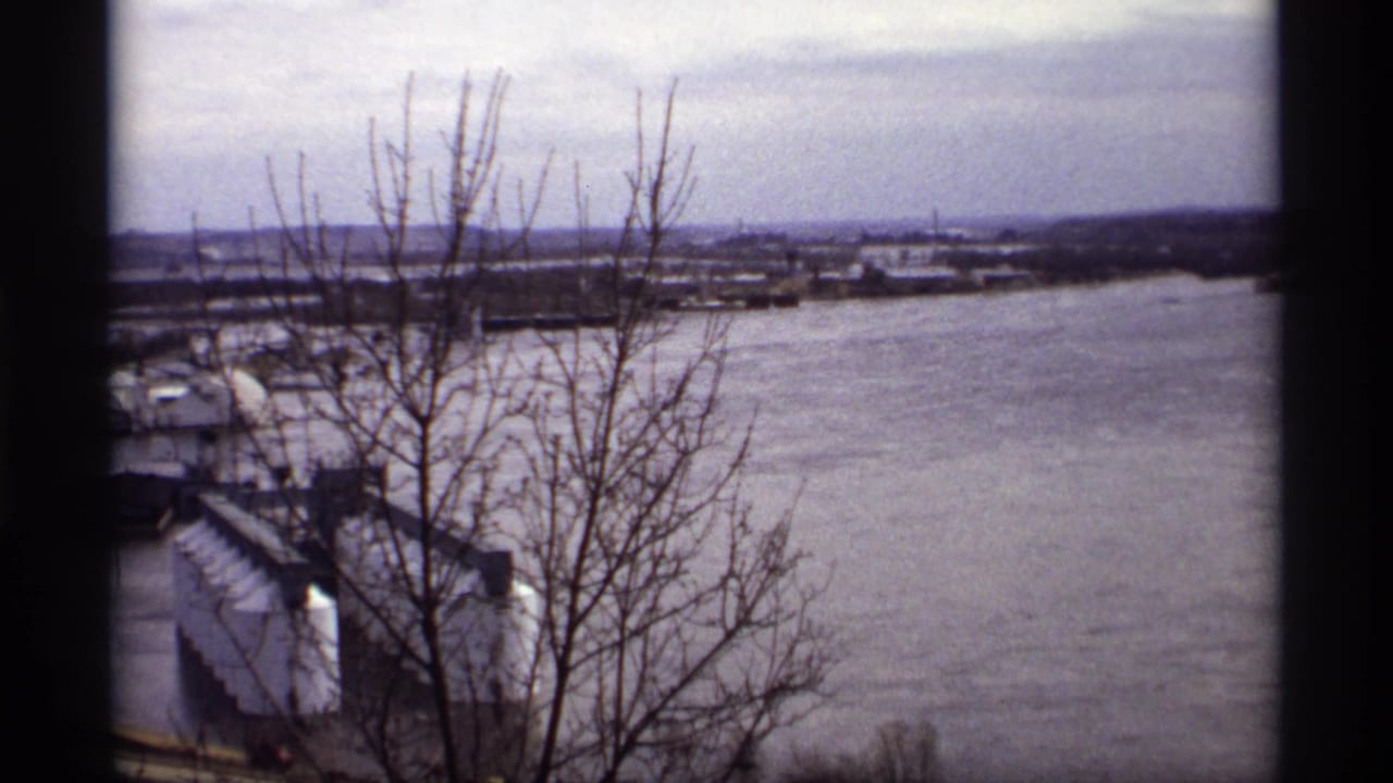 1969 Saint Paul home-movie view of floodwater around a riverfront area, with bare branches in the foreground and industrial buildings below.