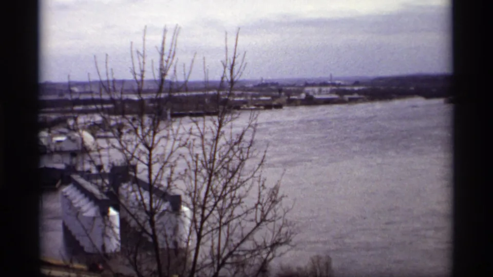 1969 Saint Paul home-movie view of floodwater around a riverfront area, with bare branches in the foreground and industrial buildings below.