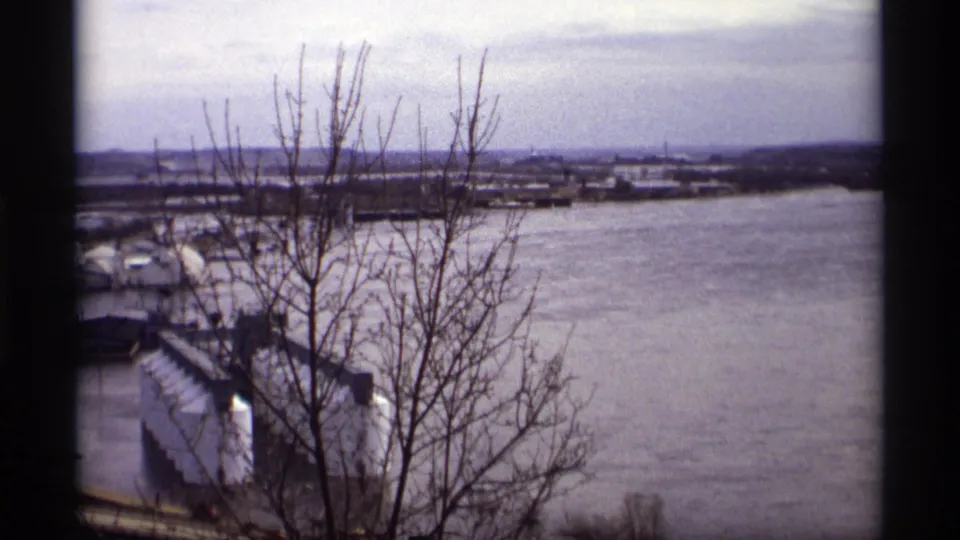Elevated 1969 Saint Paul view across a swollen river, with leafless branches in front and industrial waterfront structures partly surrounded by water.