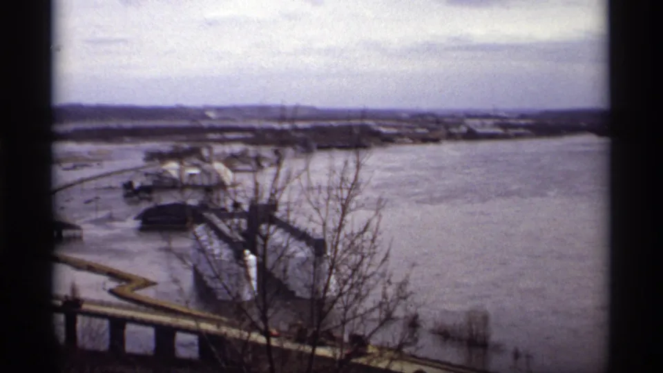 Saint Paul flood scene from 1969 showing water over low roads near industrial riverfront buildings, with bare trees and a bridge or ramp in the foreground.