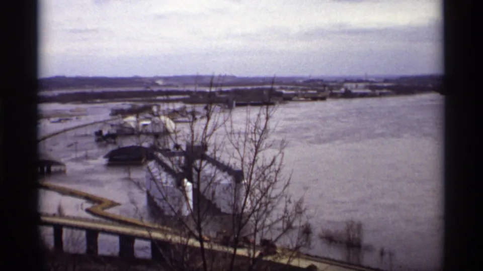 1969 Saint Paul elevated home-movie frame showing floodwater around a riverbank, bare branches in the foreground, and low industrial structures near the water.