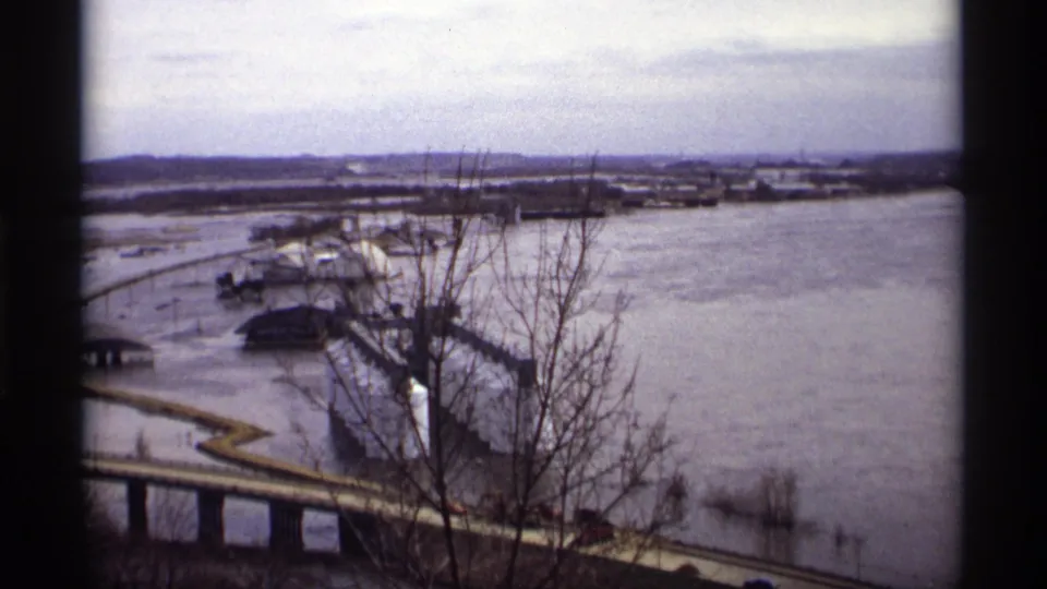 1969 Saint Paul home-movie frame showing high floodwater around roads, buildings, and a bridge under a cloudy sky.