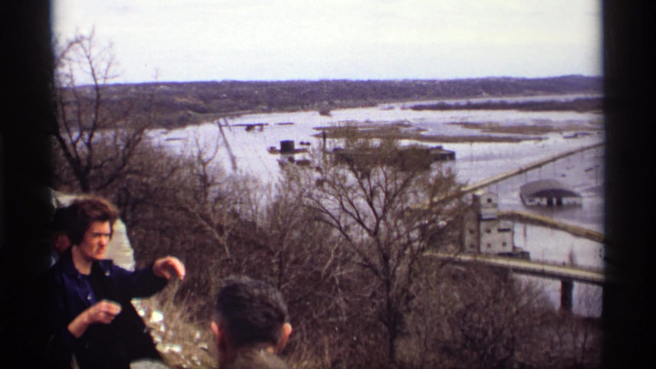 Adults at an overlook view a flooded riverfront with trees, buildings, and a bridge below.