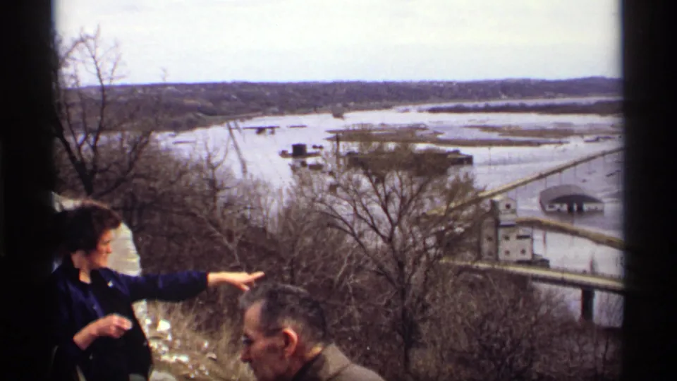 1969 Saint Paul frame with a woman pointing across flooded lowlands, with a man, bare trees, submerged roads, and buildings visible.