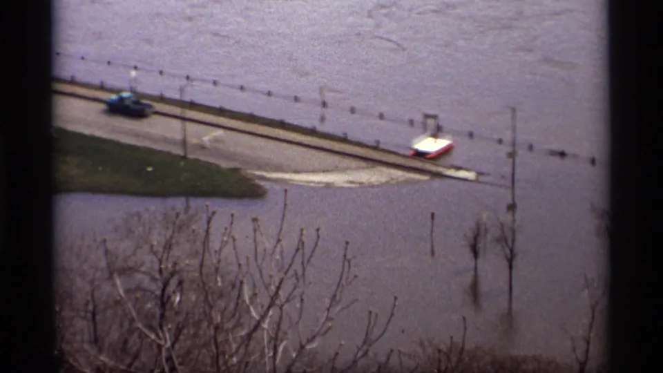 1969 Saint Paul floodwater covers a road where two vintage vehicles sit near utility poles and bare trees.