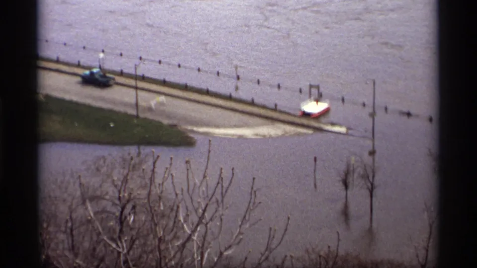 Saint Paul flood footage shows a submerged roadway with a blue car, a small white craft or vehicle, poles, and bare branches.