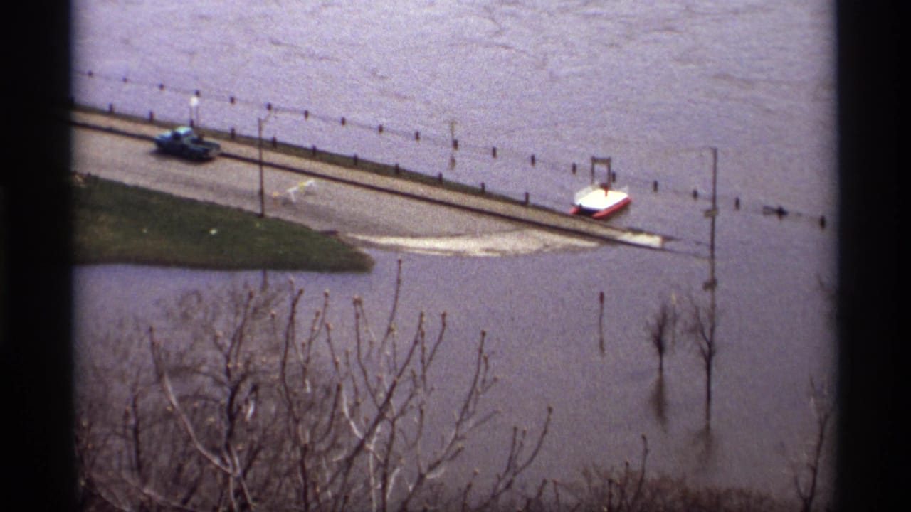 Floodwater covers a road beside a river, with a vehicle and lamp posts near the waterline.