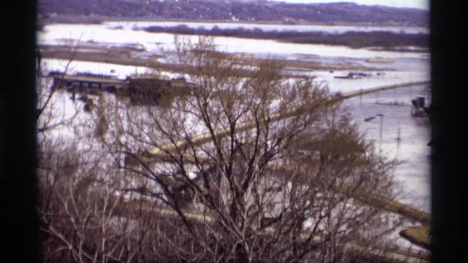 1969 Saint Paul flood scene with bare trees in front of submerged waterfront roads, a bridge or pier, and distant river flats.