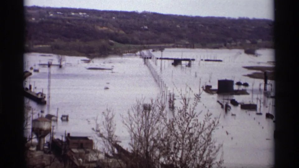 Wide 1969 Saint Paul flood view showing a long pier or causeway partly surrounded by water, bare trees, posts, and distant hillside.