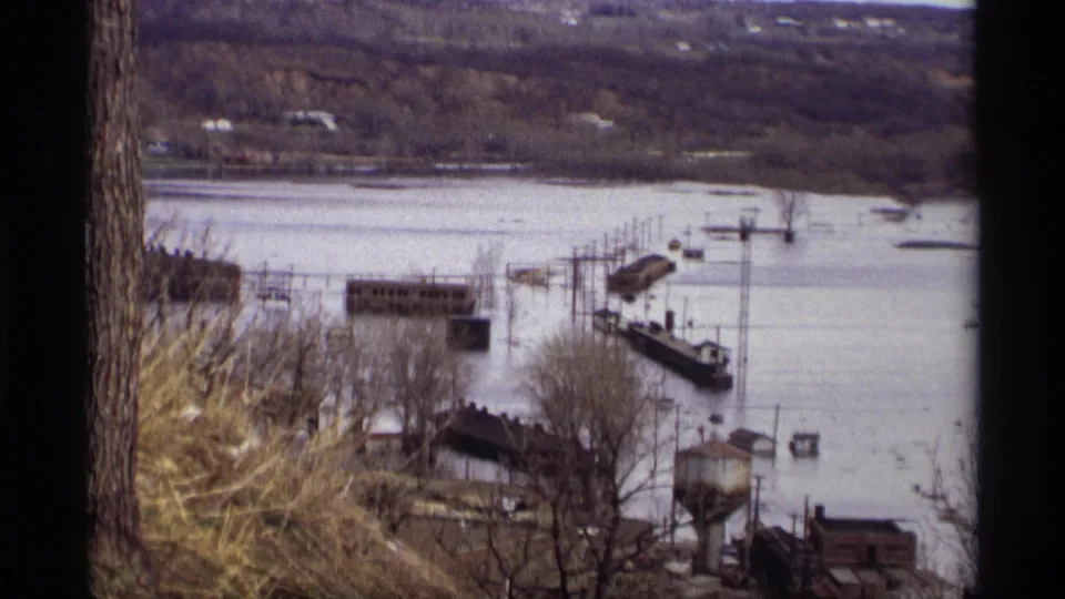 Late-1960s Saint Paul flood scene with submerged waterfront structures, bare trees, and distant hillside homes on 8mm film.