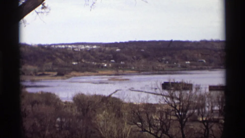 1969 8mm view of Saint Paul floodwater along a riverbank, with dock structures, leafless trees, and homes on the far slope.