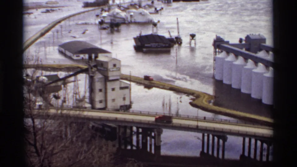Wide 1969 Saint Paul view of floodwater around riverside industrial buildings, silos, a bridge, and a red vehicle.