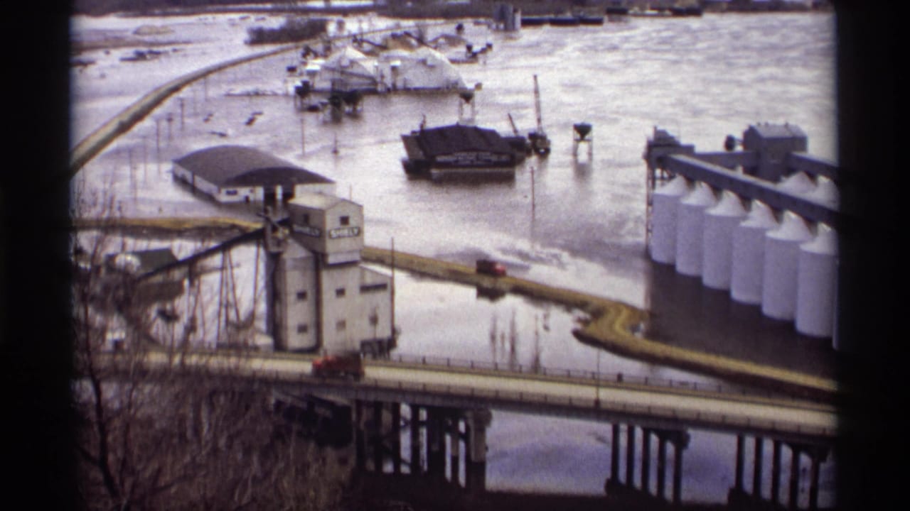 1969 Saint Paul industrial waterfront view with floodwater around a bridge, SHELBY grain elevator, white storage tanks, and barges.