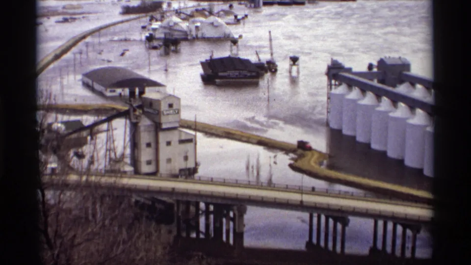 A red truck crosses a Saint Paul bridge beside floodwater, a SHELBY-marked grain elevator, barges, and white industrial tanks.