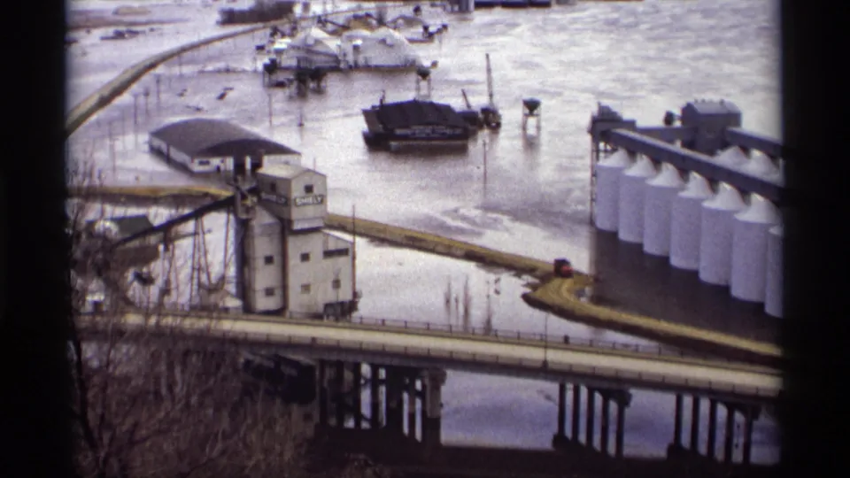 Saint Paul flood scene showing a bridge in the foreground, water around industrial buildings, white tanks, barges, and a SHELBY grain elevator.