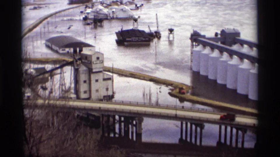 A red vehicle travels across the bridge in a 1969 Saint Paul flood view with submerged waterfront structures, barges, and white tanks.